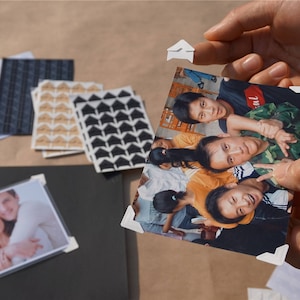 May include: A group of people, including children, are smiling and posing for a photo. The photo is held by two hands, and the corners are secured with white paper clips.