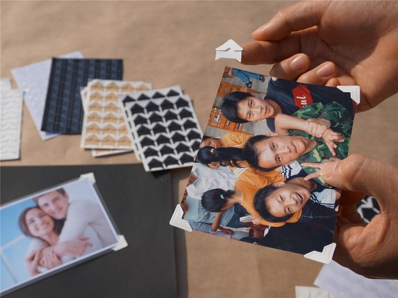 May include: A group of people, including children, are smiling and posing for a photo. The photo is held by two hands, and the corners are secured with white paper clips.