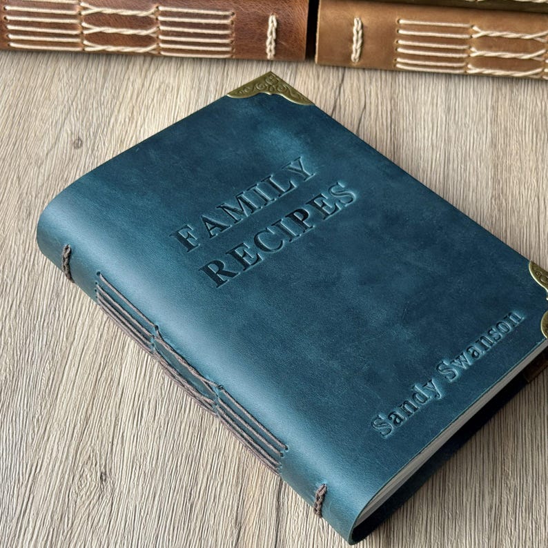 Może przedstawiać: Niebiesko-zielona sk&oacute;rzana książka kucharska z wytłoczonymi słowami "FAMILY RECIPES" na okładce. Imię "Sandy Swanson" jest r&oacute;wnież wytłoczone. Książka ma szyte wiązanie i mosiężne ochraniacze narożnik&oacute;w. Tło to drewniana powierzchnia.