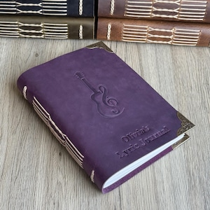 May include: A purple leather journal with a guitar and treble clef embossed on the cover. The journal has decorative corner pieces and the text "Olivia's Lyric Journal". The journal is bound with white stitching.