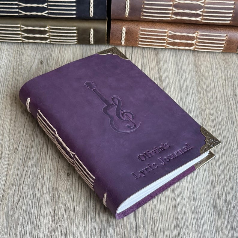 May include: A purple leather journal with a guitar and treble clef embossed on the cover. The journal has decorative corner pieces and the text "Olivia's Lyric Journal". The journal is bound with white stitching.