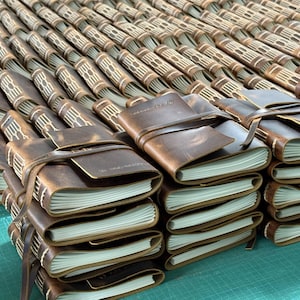 May include: A collection of brown leather journals with cream-colored pages. Each journal is bound with a leather strap closure. The journals are stacked and arranged in rows, showcasing a rustic and handcrafted aesthetic.