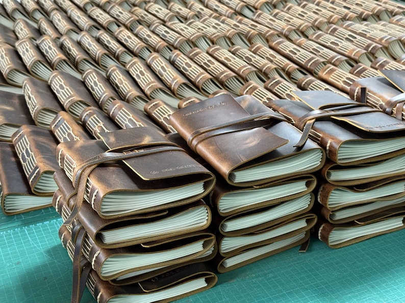 May include: A collection of brown leather journals with cream-colored pages. Each journal is bound with a leather strap closure. The journals are stacked and arranged in rows, showcasing a rustic and handcrafted aesthetic.