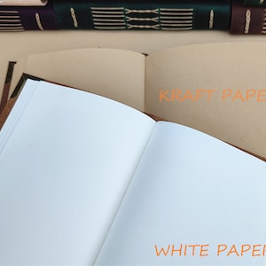 May include: Open leather-bound journal with white paper pages and a kraft paper page. The text "WHITE PAPER" and "KRAFT PAPER" are visible. Other journals are visible in the background.