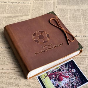 Op de afbeelding: Een vierkant bruin lederen fotoalbum met een in reliëf gemaakte voetbalafbeelding en de tekst "Ethan's Football Pilgrimage" op de kaft. Het album is voorzien van decoratieve metalen hoeken en een bruine leren sluiting aan de zijkant.