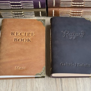 Może przedstawiać: Dwie sk&oacute;rzane książki z przepisami, jedna brązowa i jedna granatowa, ze złotymi narożnikami. Brązowa książka ma napis "RECIPE BOOK" i "Mom". Granatowa książka ma napis "Recipes" i "Gabriel Balulu".