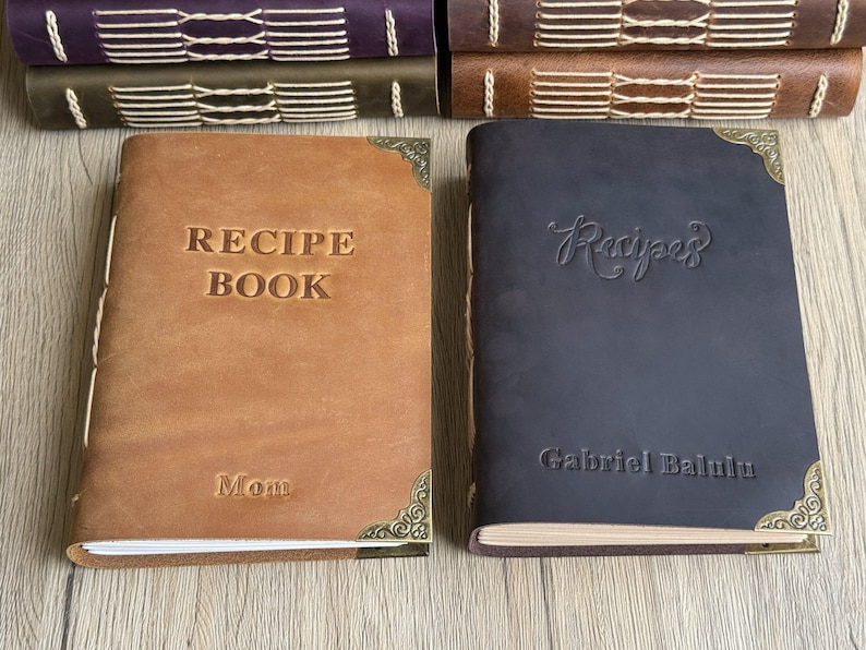 Może przedstawiać: Dwie sk&oacute;rzane książki z przepisami, jedna brązowa i jedna granatowa, ze złotymi narożnikami. Brązowa książka ma napis "RECIPE BOOK" i "Mom". Granatowa książka ma napis "Recipes" i "Gabriel Balulu".