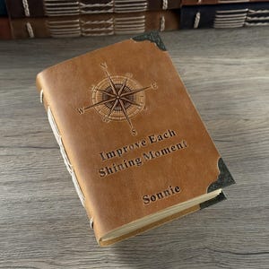 May include: A brown leather journal featuring a compass rose design and the words "Improve Each Shining Moment" and "Sonnie" embossed on the cover. The journal has metal corner protectors and is bound with white thread.