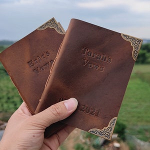 Puede incluir: Dos diarios de cuero marr&oacute;n con detalles de esquina dorados. Las cubiertas est&aacute;n grabadas con el texto "Eric's Vows" y "Tarah's Vows" y la fecha "08.2021".