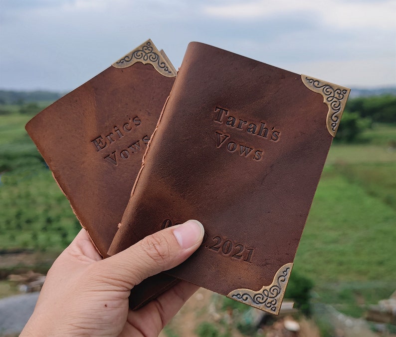 Puede incluir: Dos diarios de cuero marr&oacute;n con detalles de esquina dorados. Las cubiertas est&aacute;n grabadas con el texto "Eric's Vows" y "Tarah's Vows" y la fecha "08.2021".