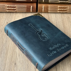 May include: A dark teal leather journal with a guitar and treble clef embossed on the cover. The journal is hand-stitched with brown thread and has the text "Emily's Lyric Journal" embossed on the cover. Brass corner protectors are visible.