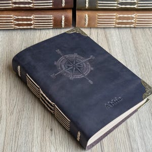 Puede incluir: Un diario de cuero azul oscuro con un diseño de rosa de los vientos en relieve en la portada. El diario tiene protectores de esquina decorativos y está cosido a mano con hilo color crema. El nombre "Abbie" está grabado en la portada.