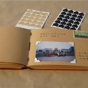 May include: A brown leather-bound scrapbook with white pages. The scrapbook has a photo of a group of people standing in front of a building. The scrapbook also has a few decorative corner stickers.