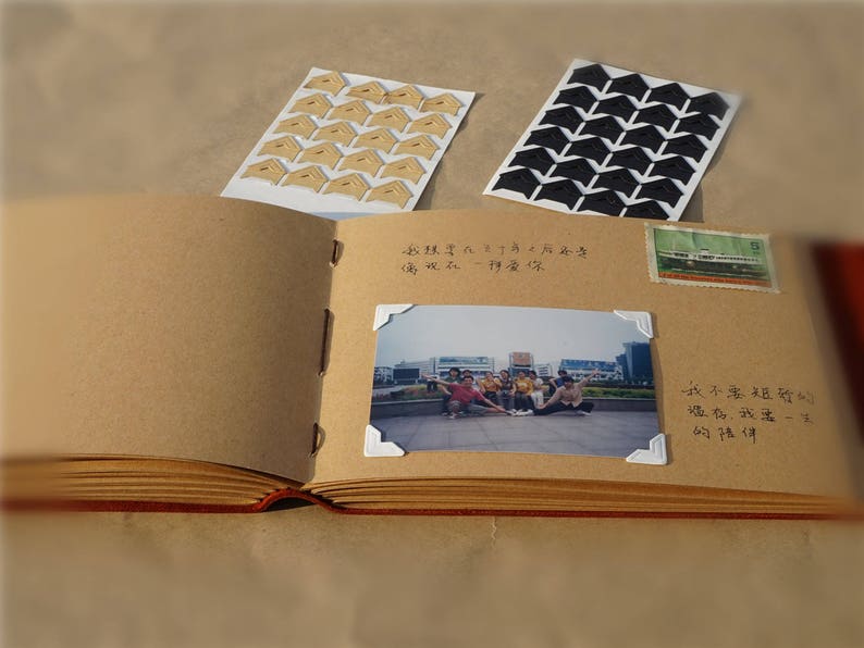 May include: A brown leather-bound scrapbook with white pages. The scrapbook has a photo of a group of people standing in front of a building. The scrapbook also has a few decorative corner stickers.