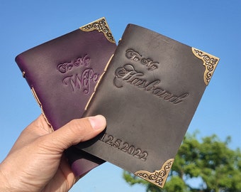May include: Two leather journals with brass-toned metal corner protectors. One journal is dark purple with the embossed text "To My Wife". The other is dark brown with the embossed text "To My Husband" and the date "12.5.2022" visible on the cover.