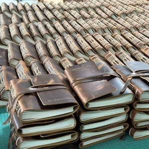 May include: A collection of brown leather journals with white-paged interiors. Each journal is bound with a leather strap closure. The journals are stacked and arranged in rows, showcasing the craftsmanship and design of the notebooks.