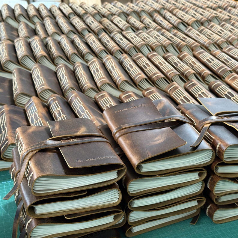 May include: A collection of brown leather journals with white-paged interiors. Each journal is bound with a leather strap closure. The journals are stacked and arranged in rows, showcasing the craftsmanship and design of the notebooks.