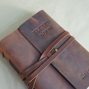 May include: A brown leather recipe book with the words "RECIPE BOOK" embossed on the cover. The book is secured with a leather strap and features the name "Kristen" embossed on the front. The book is closed.