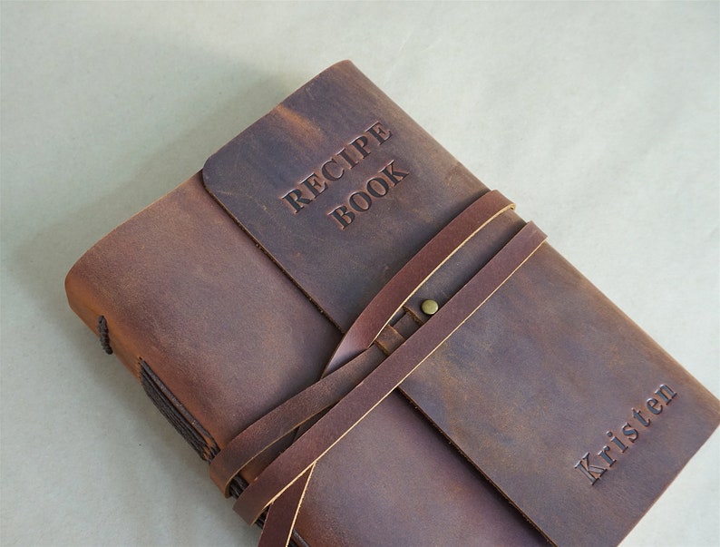 May include: A brown leather recipe book with the words "RECIPE BOOK" embossed on the cover. The book is secured with a leather strap and features the name "Kristen" embossed on the front. The book is closed.