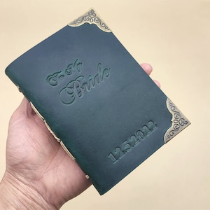 May include: A dark green leather journal with gold accents and the inscription "To My Bride 12.25.2022".