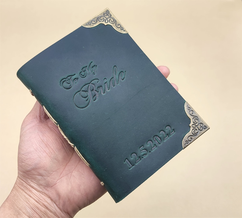 May include: A dark green leather journal with gold accents and the inscription "To My Bride 12.25.2022".