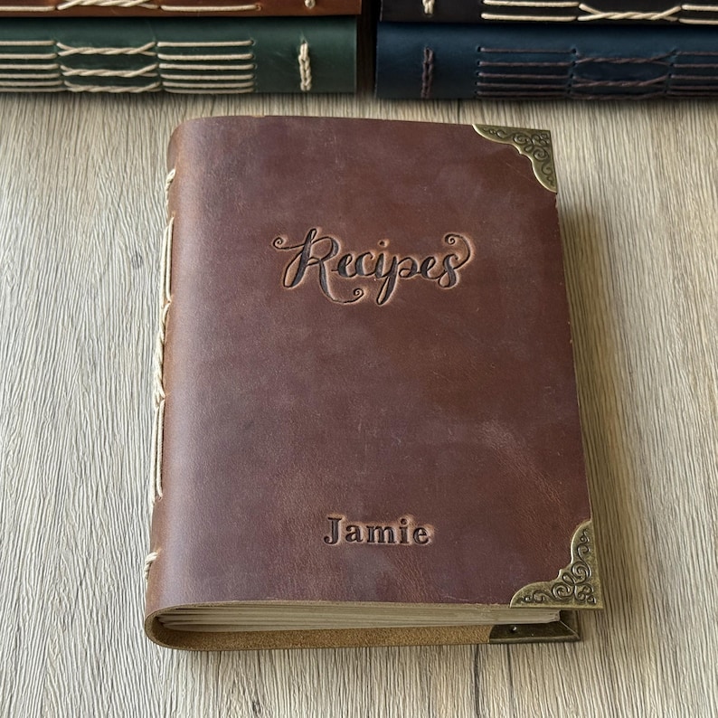 Może przedstawiać: Brązowa sk&oacute;rzana książka kucharska z wytłoczonym słowem "Recipes" na okładce. Imię "Jamie" jest r&oacute;wnież wytłoczone na okładce. Książka ma ozdobne narożniki i szyty grzbiet. Inne książki są widoczne w tle.
