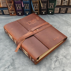 May include: A brown leather-bound journal with a strap closure. The journal has the word "Recipes" written on the front cover in cursive script and the name "Mayra" written on the back cover.