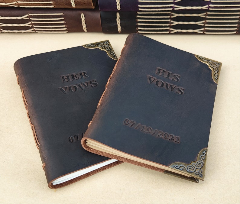 Puede incluir: Dos diarios de cuero marr&oacute;n oscuro con esquinas de metal dorado. Las tapas est&aacute;n grabadas con las palabras "Her Vows" y "His Vows" y la fecha "07/19/2021".