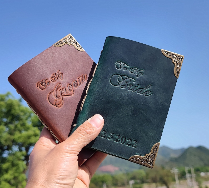 May include: Two leather-bound journals with ornate brass corners. The brown journal says "To My Groom" and the green journal says "To My Bride" with the date "12.25.2022".