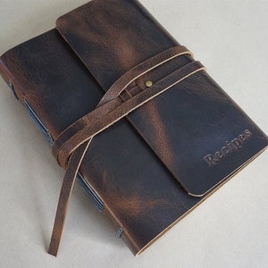 May include: A dark brown leather recipe journal with a wrap-around strap. The word "Recipes" is embossed on the cover. The journal has a rustic, vintage aesthetic.