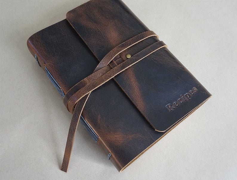 May include: A dark brown leather recipe journal with a wrap-around strap. The word "Recipes" is embossed on the cover. The journal has a rustic, vintage aesthetic.