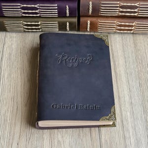 Może przedstawiać: Granatowy sk&oacute;rzany zeszyt z przepisami z wytłoczonym słowem "Recipes" na okładce. Imię "Gabriel Balulu" jest r&oacute;wnież wytłoczone na okładce. Książka ma ozdobne narożniki i leży na drewnianej powierzchni.