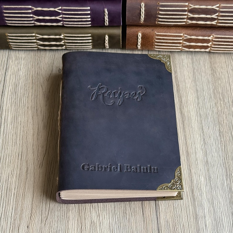 Może przedstawiać: Granatowy sk&oacute;rzany zeszyt z przepisami z wytłoczonym słowem "Recipes" na okładce. Imię "Gabriel Balulu" jest r&oacute;wnież wytłoczone na okładce. Książka ma ozdobne narożniki i leży na drewnianej powierzchni.