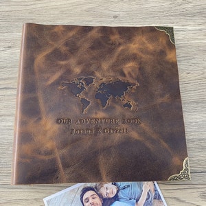 Może przedstawiać: Brązowy skórzany album na zdjęcia z wytłoczonym na froncie mapą świata. Album nosi tytuł "Our Adventure Book" z nazwiskami "Joshua & Garrett" poniżej.