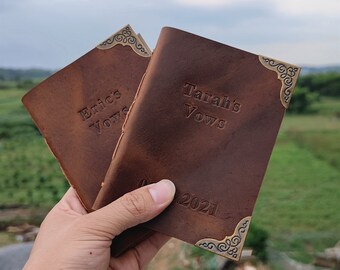 May include: Two brown leather journals with gold corner accents. The covers are embossed with the text "Eric's Vows" and "Tarah's Vows" and the date "08.2021".