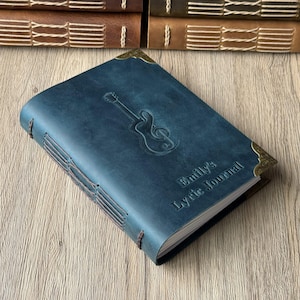 May include: A blue leather journal with a guitar and treble clef embossed on the cover. The journal is titled "Emily's Lyric Journal" and has decorative corner pieces. The binding is hand-stitched with brown thread.