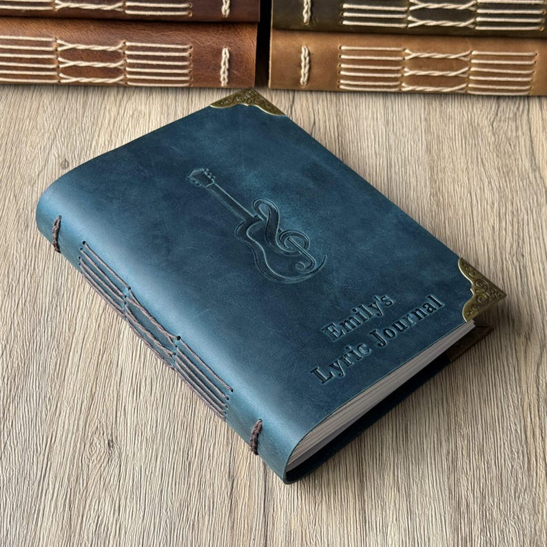 May include: A blue leather journal with a guitar and treble clef embossed on the cover. The journal is titled "Emily's Lyric Journal" and has decorative corner pieces. The binding is hand-stitched with brown thread.