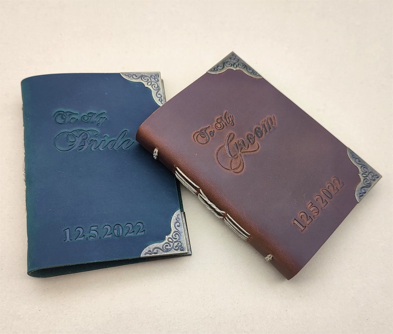May include: Two leather-bound journals with decorative metal corners. The blue journal has the text "To My Bride 12.5.2022" and the brown journal has the text "To My Groom 12.5.2022".