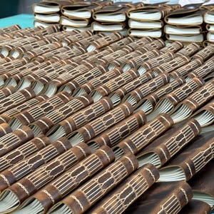 May include: A collection of brown leather journals with cream-colored pages. The journals are arranged in rows, showcasing their hand-stitched binding with white thread. The leather has a distressed look, and the pages are slightly visible.