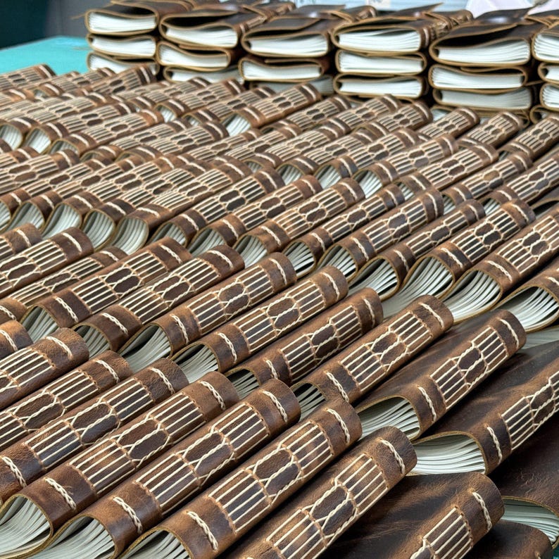 May include: A collection of brown leather journals with cream-colored pages. The journals are arranged in rows, showcasing their hand-stitched binding with white thread. The leather has a distressed look, and the pages are slightly visible.