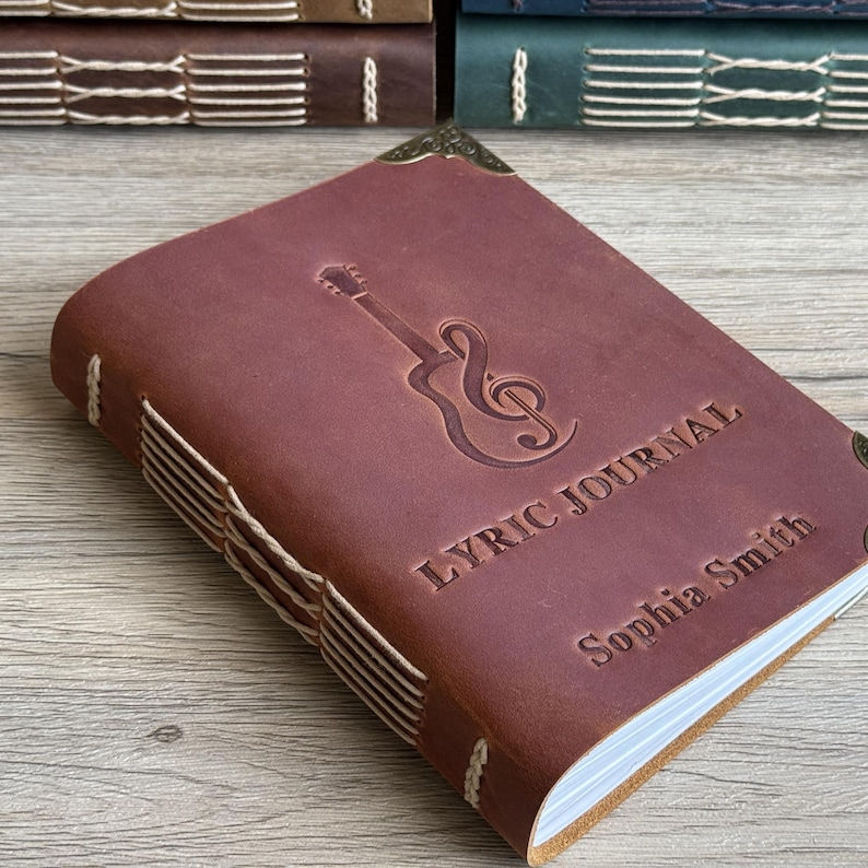 May include: A brown leather Lyric Journal with a guitar and treble clef design. The journal is personalized with the name "Sophia Smith". The journal has decorative corner pieces and is bound with white stitching.