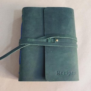 May include: A teal leather recipe journal with a wrap-around strap closure. The word "Recipes" is embossed on the front cover. Blue stitching is visible along the spine. The journal is closed with a leather strap and a brass button.