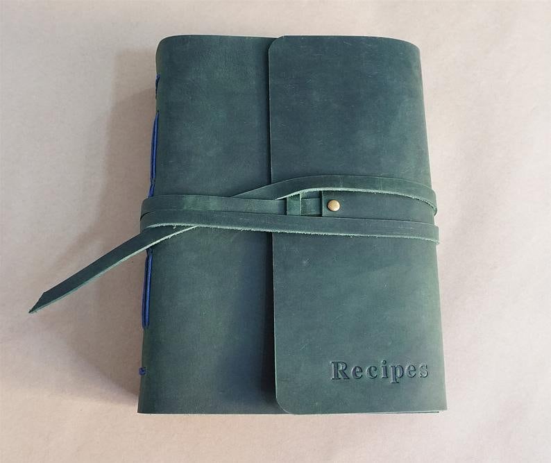 May include: A teal leather recipe journal with a wrap-around strap closure. The word "Recipes" is embossed on the front cover. Blue stitching is visible along the spine. The journal is closed with a leather strap and a brass button.