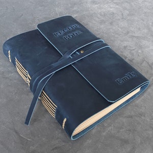 May include: A dark blue leather-bound recipe book with a strap closure and the words "Recipe Book" embossed on the front cover. The book is personalized with the name "Emily" on the back cover.