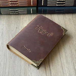 Może przedstawiać: Brązowa sk&oacute;rzana książka kucharska z wytłoczonym napisem "Recipes" na okładce oraz imieniem "Jamie". Książka ma ozdobne narożniki i widoczne kremowe strony. Inne książki w sk&oacute;rzanej oprawie są ułożone w tle.