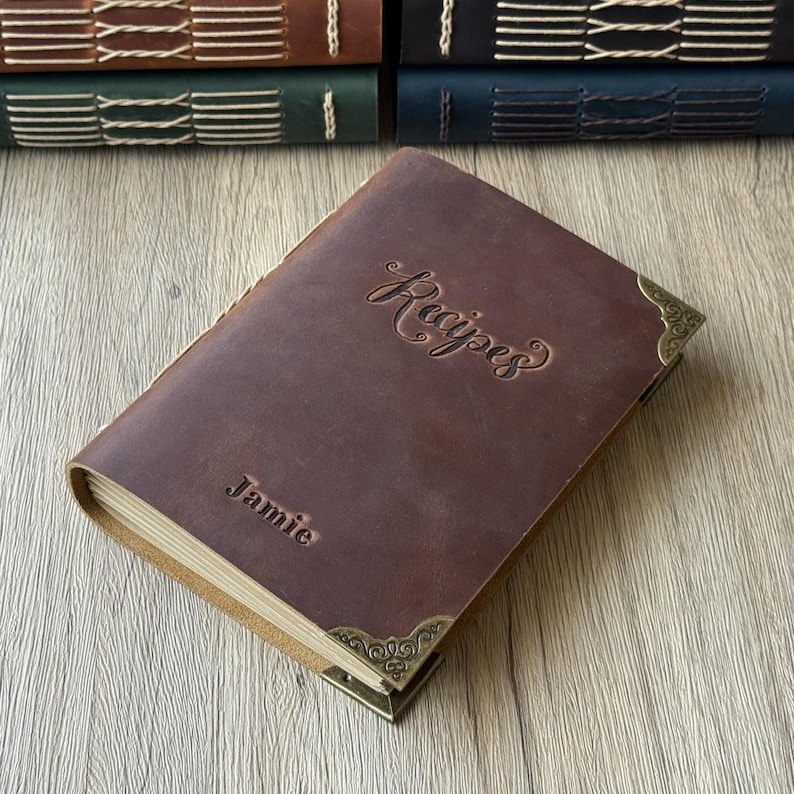 Może przedstawiać: Brązowa sk&oacute;rzana książka kucharska z wytłoczonym napisem "Recipes" na okładce oraz imieniem "Jamie". Książka ma ozdobne narożniki i widoczne kremowe strony. Inne książki w sk&oacute;rzanej oprawie są ułożone w tle.
