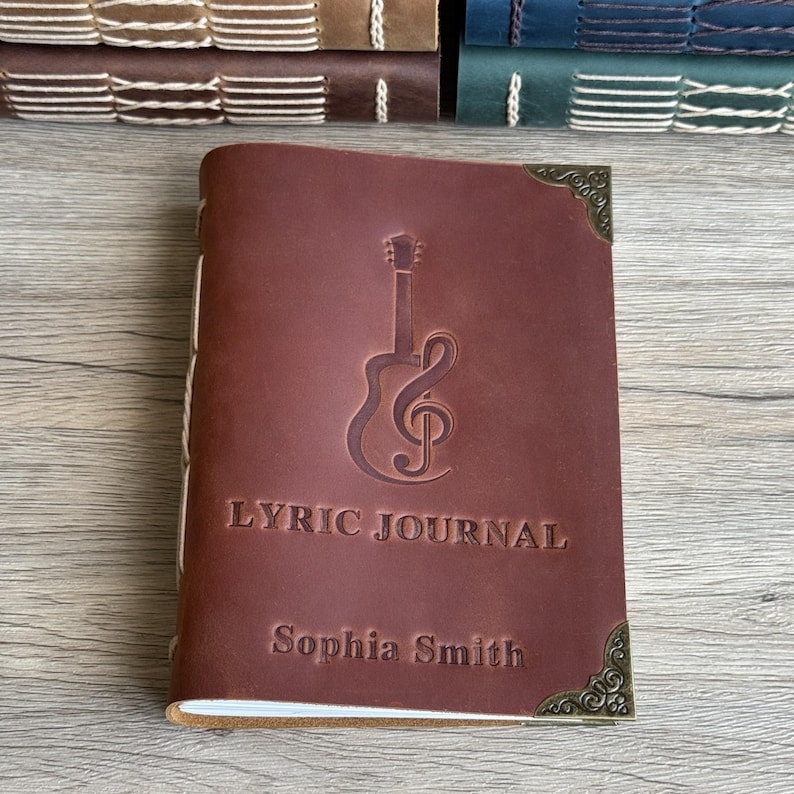 May include: A brown leather Lyric Journal with a guitar and treble clef design. The journal is embossed with the words "LYRIC JOURNAL" and "Sophia Smith". Decorative corner accents add detail. Other journals are visible in the background.