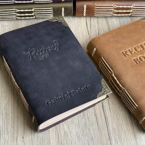Puede incluir: Dos libros de recetas encuadernados en cuero. Uno es azul oscuro con "Recipes" en relieve en la portada y el nombre "Gabriel Balint" debajo. El otro es marrón claro con "RECIPE BOOK" en relieve. Ambos tienen esquinas decorativas.