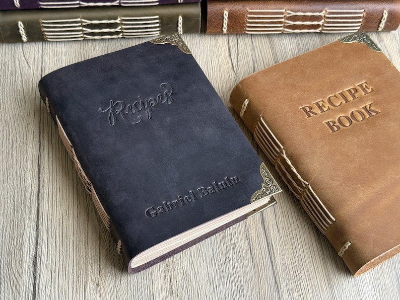 Może przedstawiać: Dwa sk&oacute;rzane książki kucharskie. Jedna jest granatowa z wytłoczonym napisem "Recipes" na okładce i nazwiskiem "Gabriel Balint" poniżej. Druga jest brązowa z wytłoczonym napisem "RECIPE BOOK". Obie mają ozdobne narożniki.