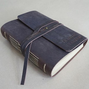 May include: A dark brown leather-bound recipe book with the words "FAMILY RECIPES" embossed on the cover. The book is secured with a leather strap and features cream-colored pages and white stitching along the spine.